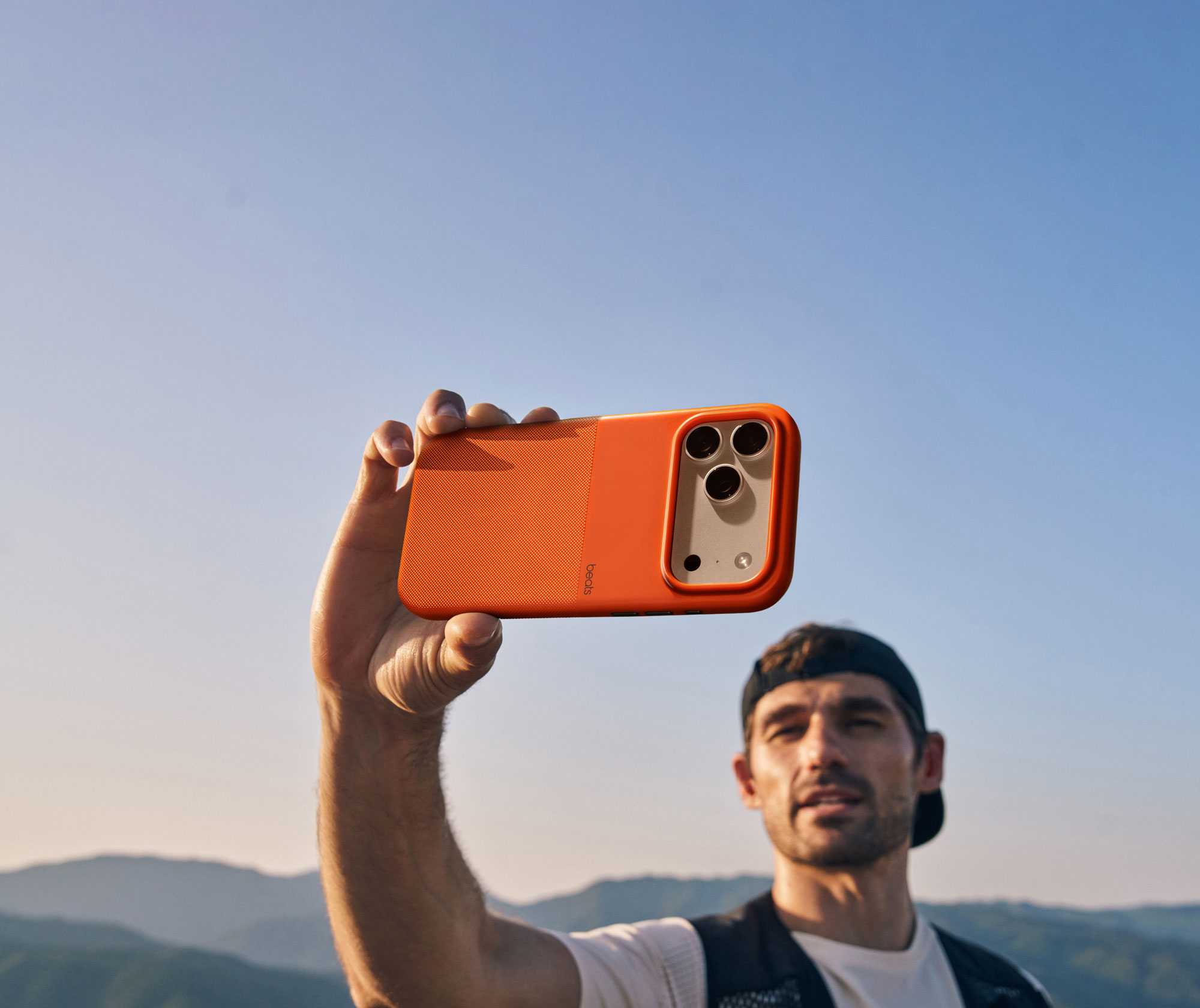 A man holding the Beats iPhone 17 rugged case with MagSafe and Camera Control in Sierra Orange, against a backdrop of mountains and a clear sky.